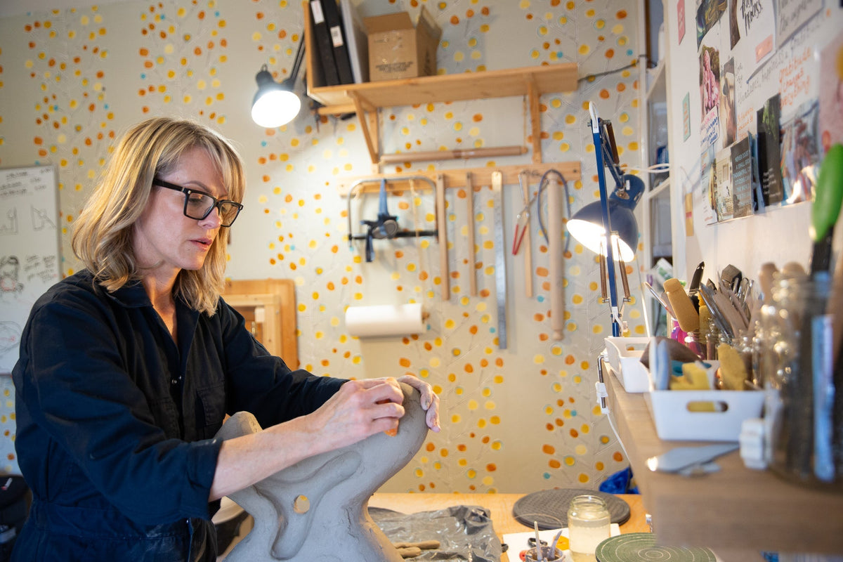 Rachel Peters working in her home ceramic sculpture studio, shaping an unglazed stoneware form surrounded by tools and materials.