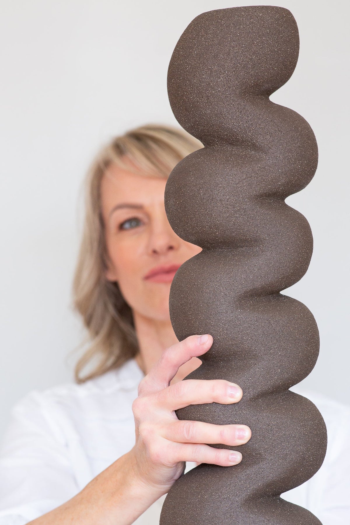 Rachel Peters touching a tall hand-built ceramic sculpture with a stacked organic form in unglazed dark stoneware, demonstrating the tactile quality of her work.