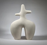 An abstract figurative ceramic sculpture with rounded legs and a branching upper torso, in a matte off-white finish on a light grey studio background.