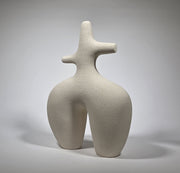 An abstract figurative ceramic sculpture with rounded legs and a branching upper torso, in a matte off-white finish on a light grey studio background.