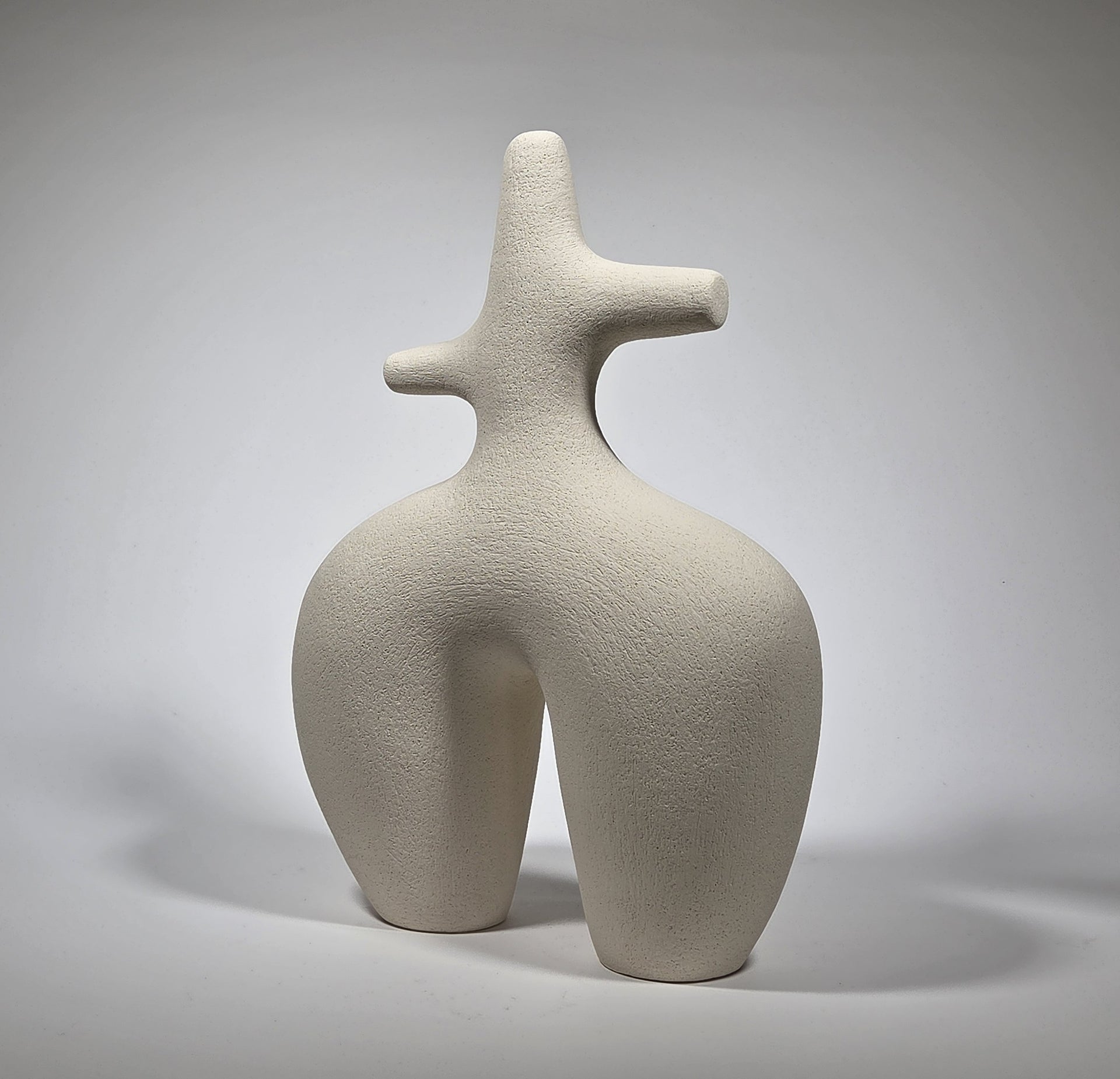 An abstract figurative ceramic sculpture with rounded legs and a branching upper torso, in a matte off-white finish on a light grey studio background.
