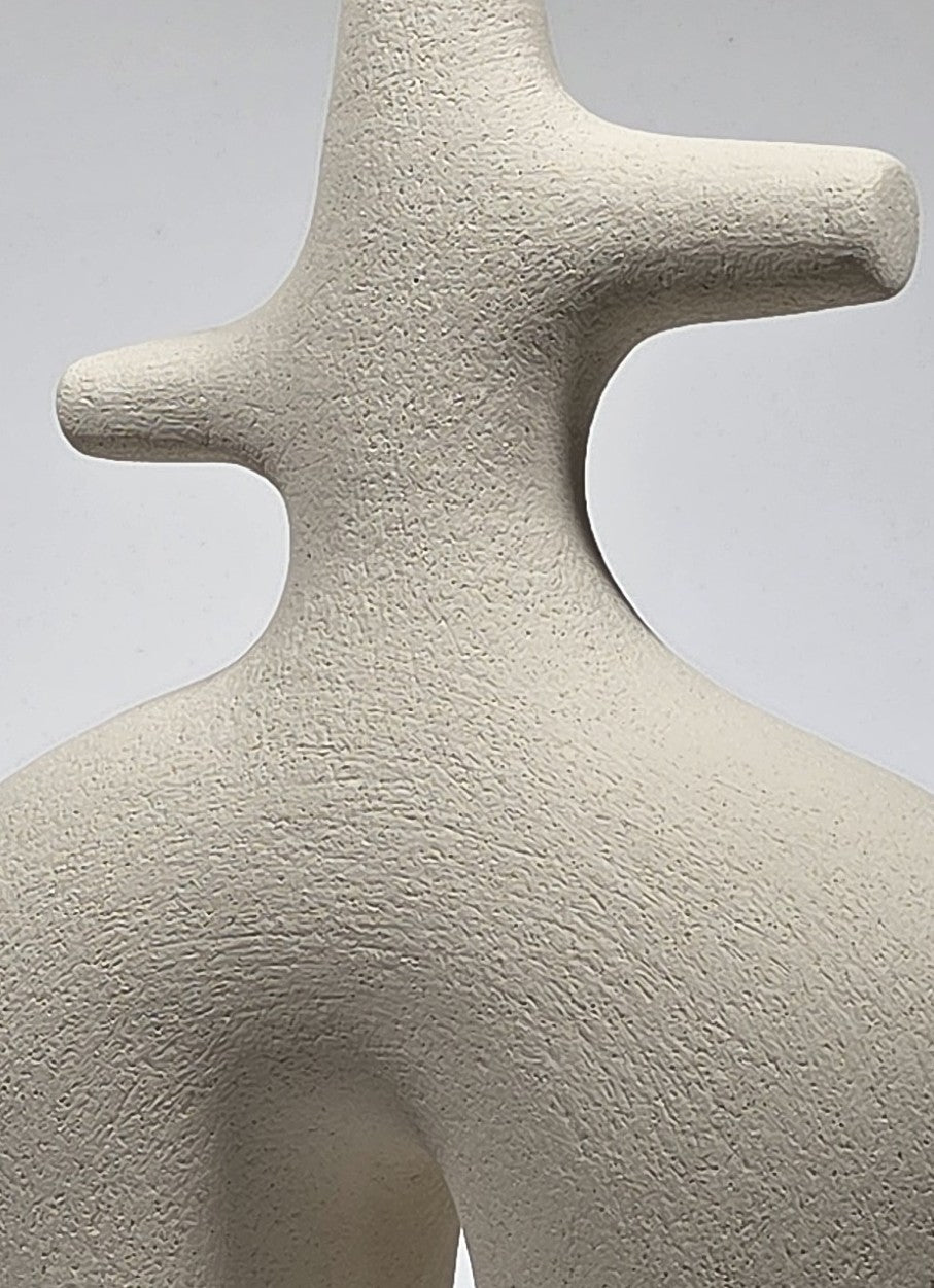 A close-up of a textured, abstract ceramic sculpture in a light warm gray or off-white color, showing the organic, cross-like form against a neutral gray background.