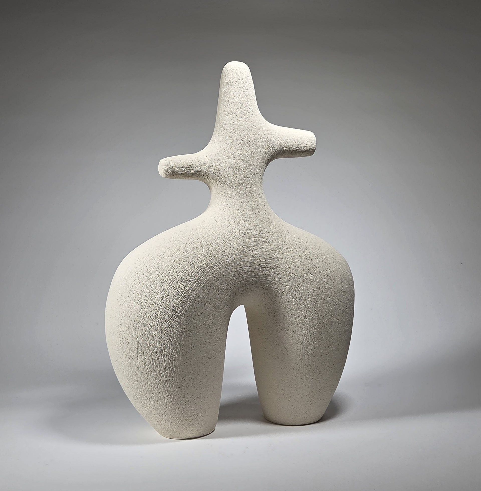 An abstract figurative ceramic sculpture with rounded legs and a branching upper torso, in a matte off-white finish on a light grey studio background.