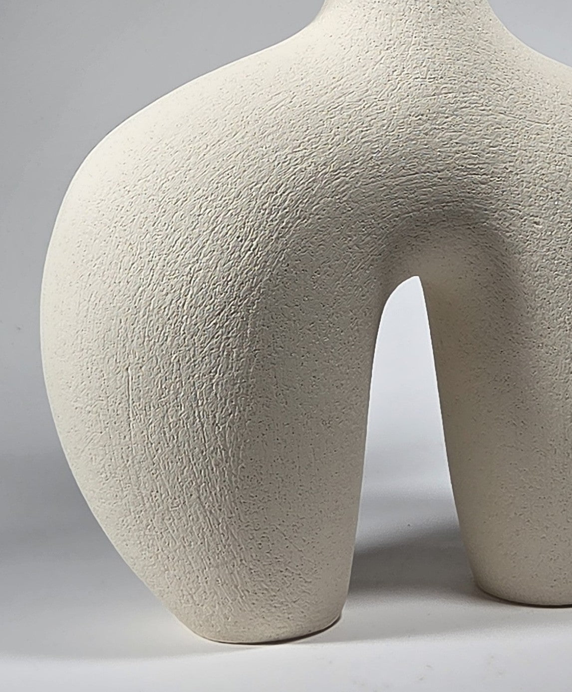 Close up of the bottom section of an abstract figurative ceramic sculpture showing rounded legs, in a matte off-white finish on a light grey studio background.