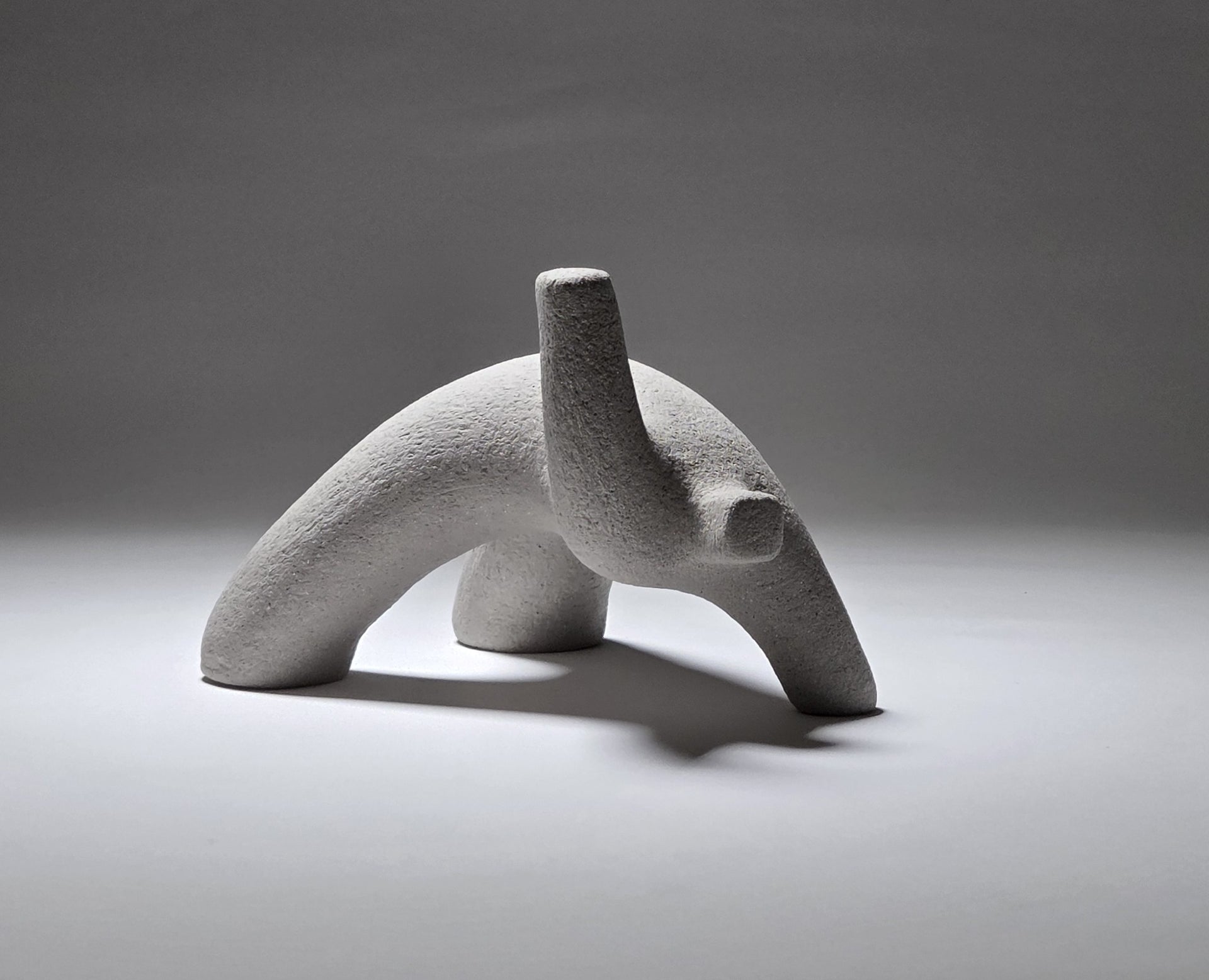 Abstract grey ceramic figure arching backwards with one arm on the floor and one stretched upwards pictures on a light grey background