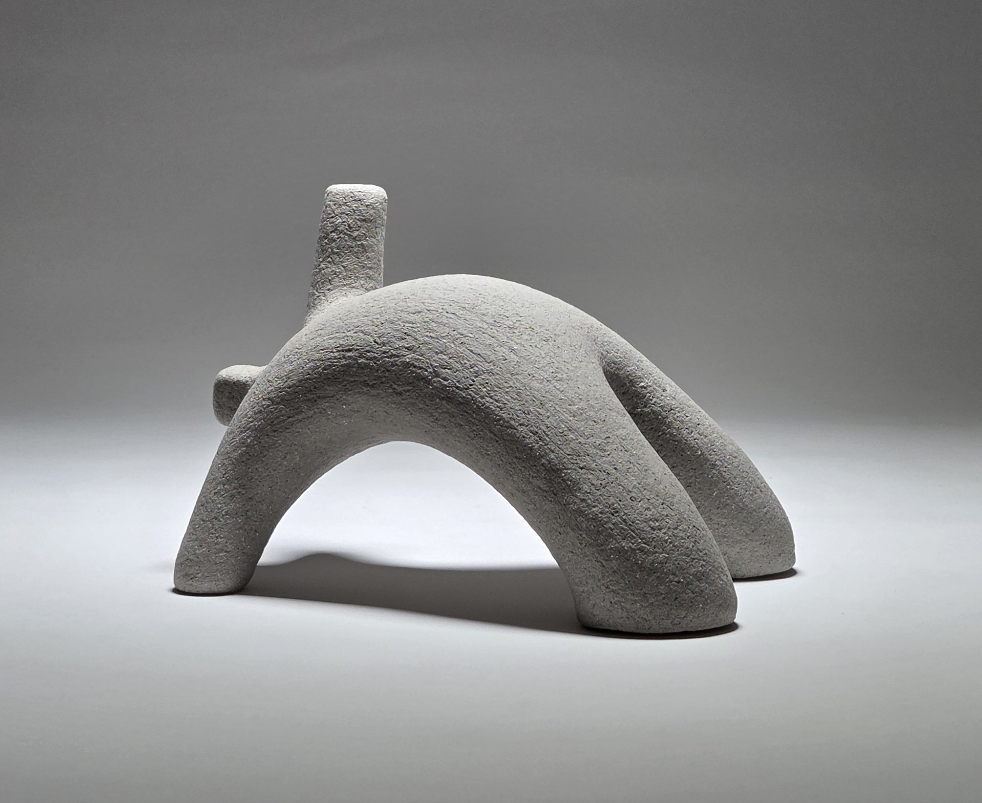 Abstract grey ceramic figure arching backwards with one arm on the floor and one stretched upwards pictures on a light grey background