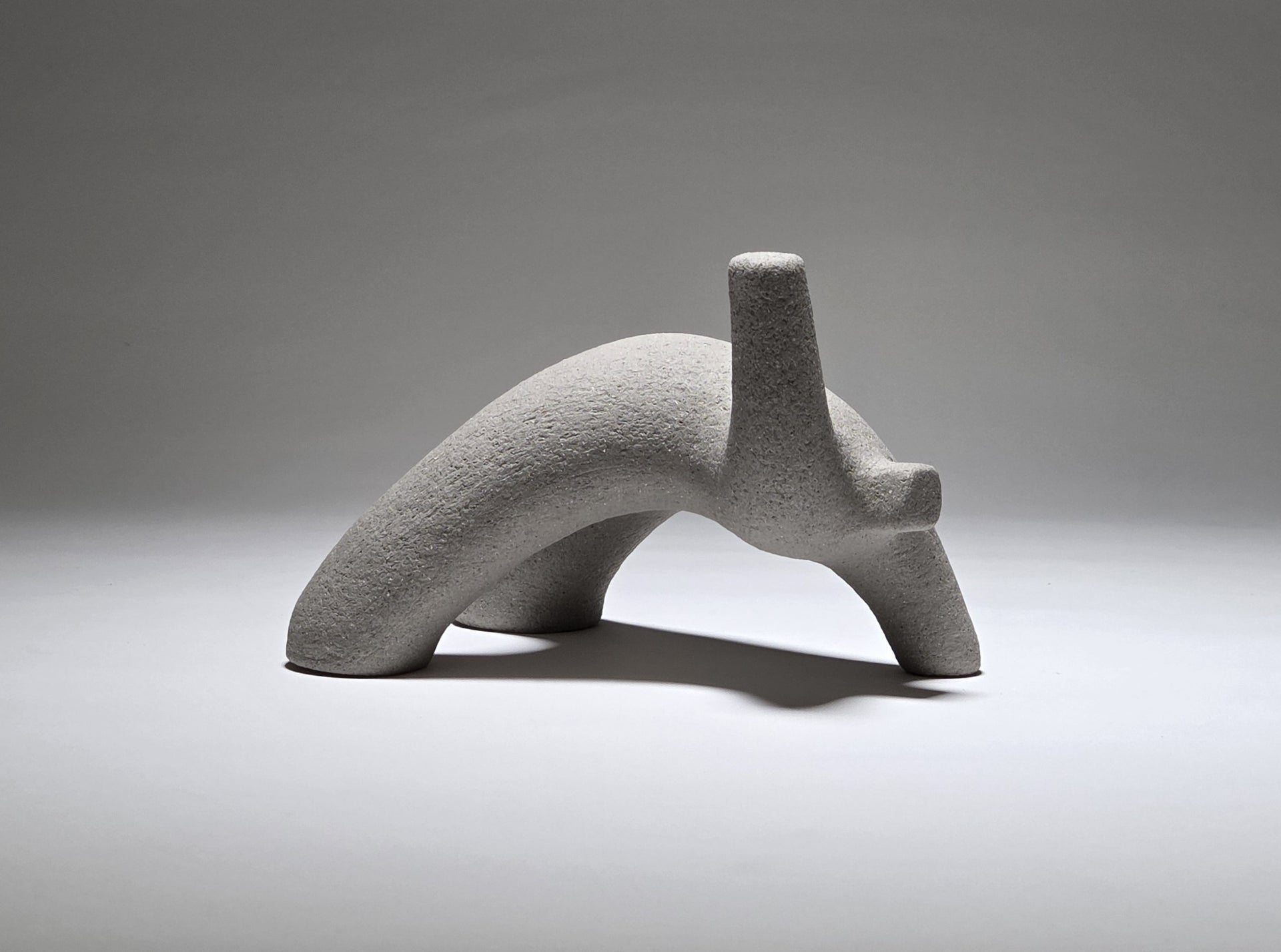 Abstract grey ceramic figure arching backwards with one arm on the floor and one stretched upwards pictures on a light grey background